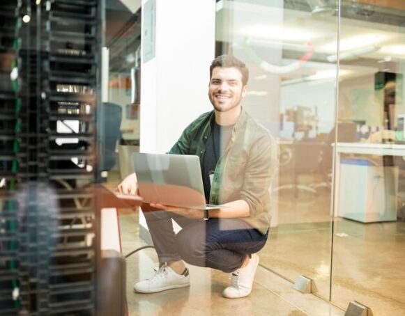 portrait-happy-young-male-network-engineer-with-laptop-hand-working-datacenter